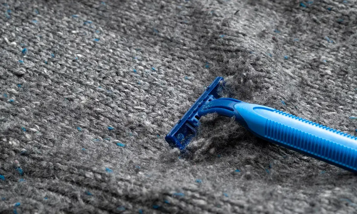 En blå rakhyvel tar bort noppor från en grå stickad tröja i polyester.