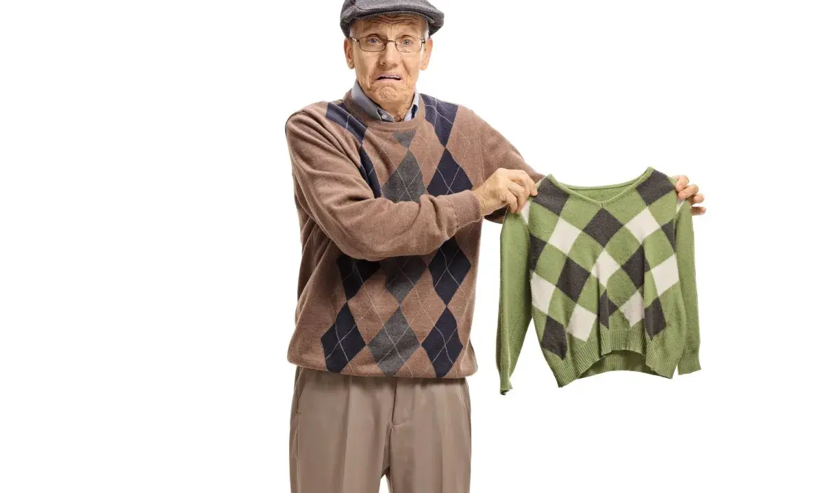 Äldre man med argyle-tröja håller upp en mindre, likadan tröja. Han ser bekymrad ut, som om bomullen krymper.