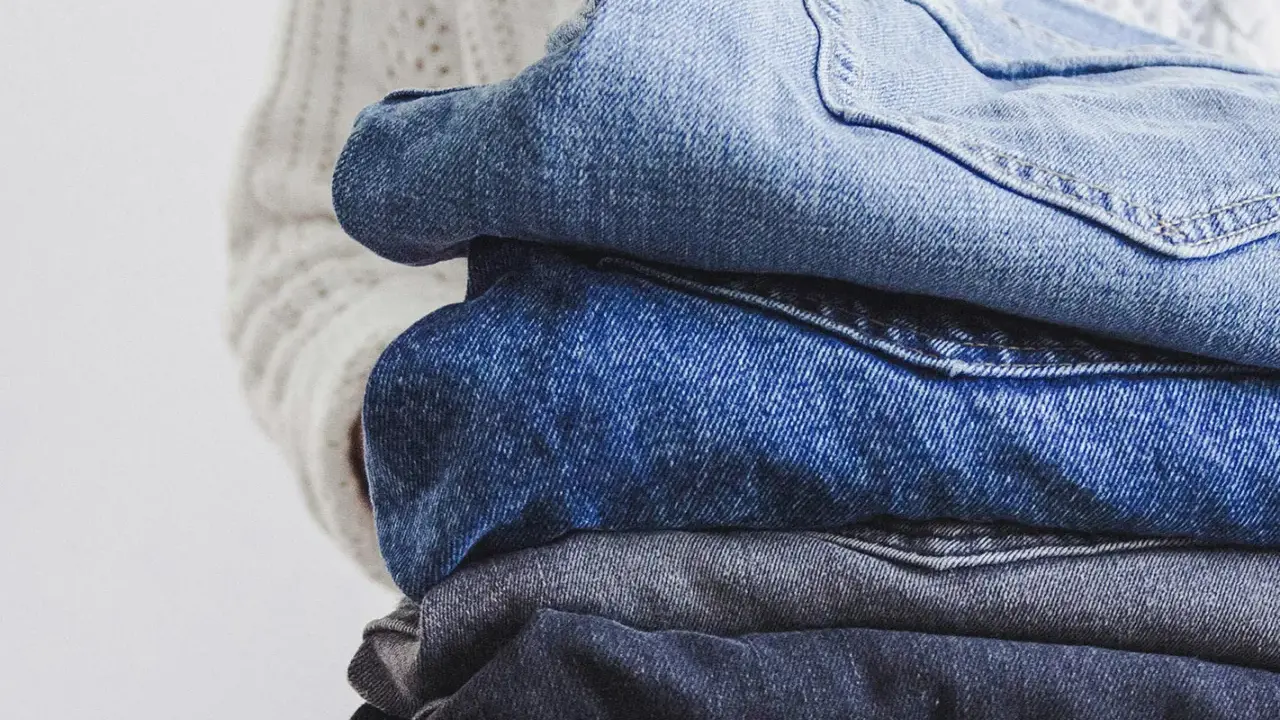 En hög med jeans i olika blå nyanser, redo för jeans återvinning. En vit stickad tröja syns i bakgrunden.