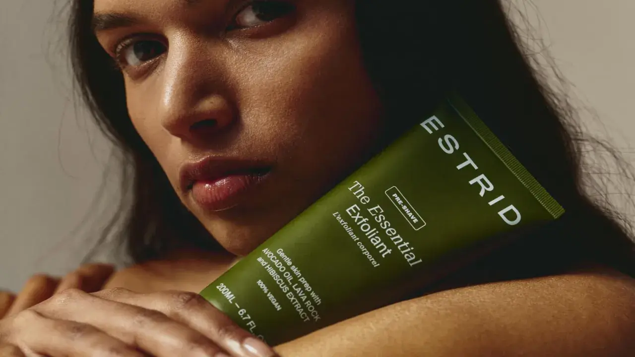 En kvinna håller en grön tub med "The Essential Exfoliant" från ESTRID. Perfekt för att lära sig hur exfolierar man huden för en mjukare känsla.