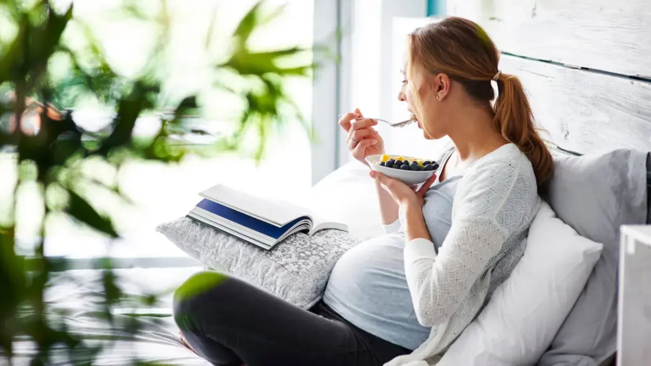 Gravid kvinna med svullen mage äter frukost i sängen. En bok ligger bredvid.