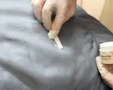 Händer med handskar applicerar filler för att laga reva i tyg. En burk med "Heavy Filler" syns.