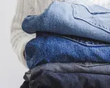 En hög med jeans i olika blå nyanser, redo för jeans återvinning. En vit stickad tröja syns i bakgrunden.