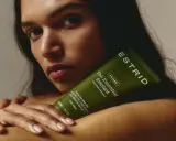 En kvinna håller en grön tub med "The Essential Exfoliant" från ESTRID. Perfekt för att lära sig hur exfolierar man huden för en mjukare känsla.