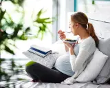 Gravid kvinna med svullen mage äter frukost i sängen. En bok ligger bredvid.