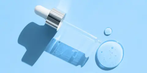 En pipett med klar vätska, som innehåller hyaluronsyra och retinol, ligger på en ljusblå bakgrund med droppar av serum.