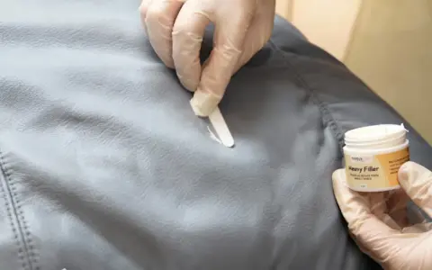 Händer med handskar applicerar filler för att laga reva i tyg. En burk med "Heavy Filler" syns.