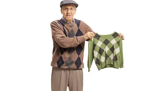Äldre man med argyle-tröja håller upp en mindre, likadan tröja. Han ser bekymrad ut, som om bomullen krymper.