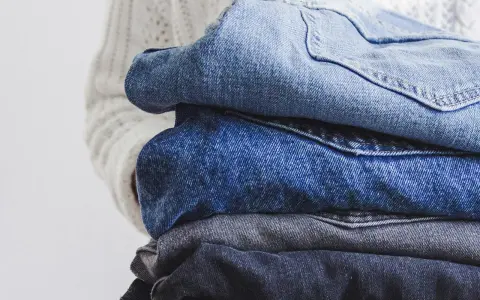En hög med jeans i olika blå nyanser, redo för jeans återvinning. En vit stickad tröja syns i bakgrunden.