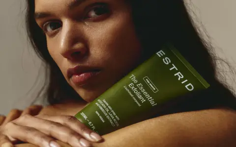 En kvinna håller en grön tub med "The Essential Exfoliant" från ESTRID. Perfekt för att lära sig hur exfolierar man huden för en mjukare känsla.