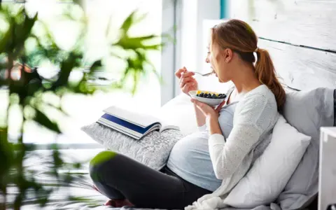 Gravid kvinna med svullen mage äter frukost i sängen. En bok ligger bredvid.