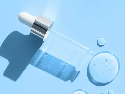 En pipett med klar vätska, som innehåller hyaluronsyra och retinol, ligger på en ljusblå bakgrund med droppar av serum.