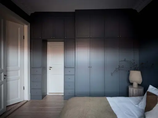 Mörka garderober i stockholm stil ramar in en vit dörr. En vas med grenar står på ett sidobord bredvid en säng.