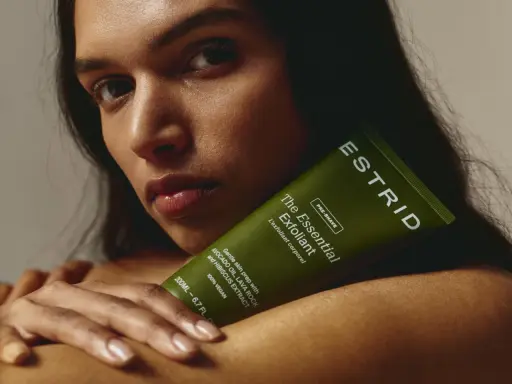 En kvinna håller en grön tub med "The Essential Exfoliant" från ESTRID. Perfekt för att lära sig hur exfolierar man huden för en mjukare känsla.