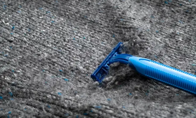 En blå rakhyvel tar bort noppor från en grå stickad tröja i polyester.