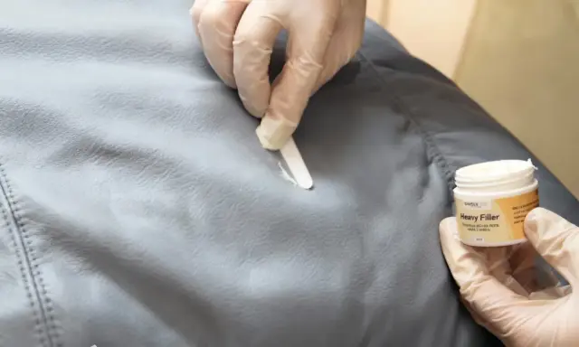 Händer med handskar applicerar filler för att laga reva i tyg. En burk med "Heavy Filler" syns.