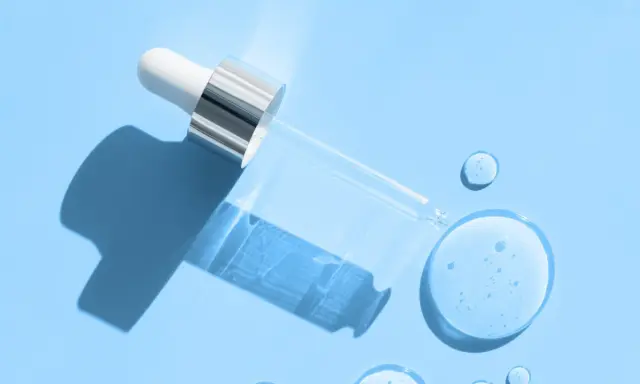 En pipett med klar vätska, som innehåller hyaluronsyra och retinol, ligger på en ljusblå bakgrund med droppar av serum.