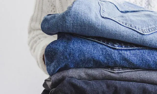 En hög med jeans i olika blå nyanser, redo för jeans återvinning. En vit stickad tröja syns i bakgrunden.