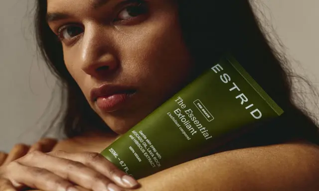 En kvinna håller en grön tub med "The Essential Exfoliant" från ESTRID. Perfekt för att lära sig hur exfolierar man huden för en mjukare känsla.
