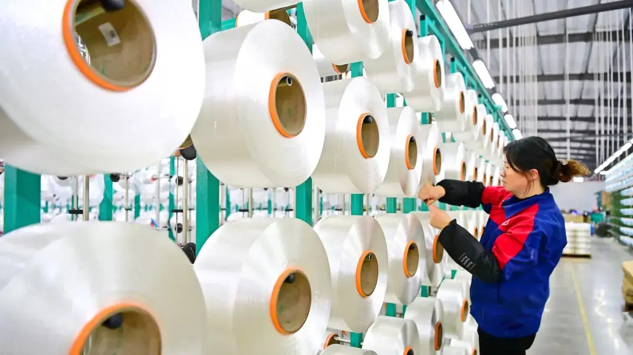 En kvinna arbetar i en fabrik där stora rullar med garn hänger på ställningar. Detta är en del av hur tillverkas polyester, en process som ger oss många av våra textilier.