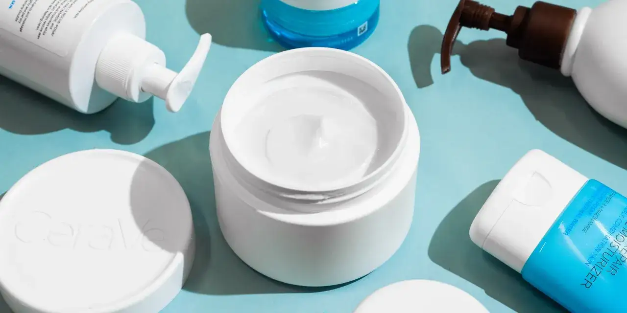 En &ouml;ppen burk med vit kr&auml;m, perfekt f&ouml;r att applicera body lotion i ansiktet. Flera hudv&aring;rdsprodukter ligger utspridda.
