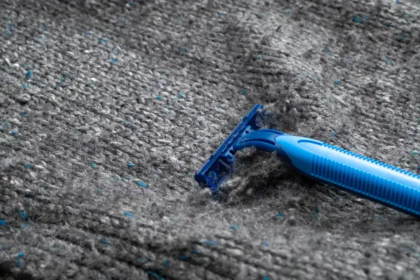 En blå rakhyvel tar bort noppor från en grå stickad tröja i polyester.