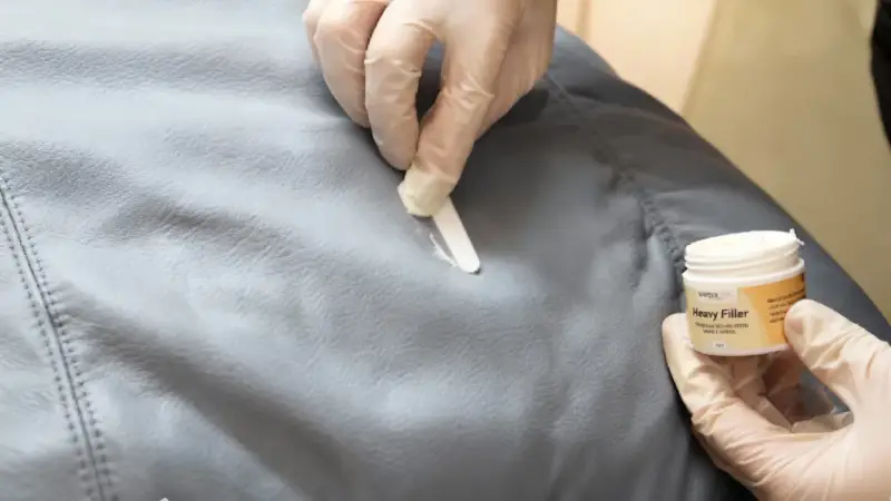 Händer med handskar applicerar filler för att laga reva i tyg. En burk med "Heavy Filler" syns.