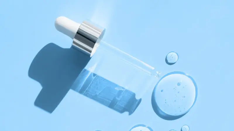 En pipett med klar vätska, som innehåller hyaluronsyra och retinol, ligger på en ljusblå bakgrund med droppar av serum.